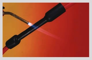 A black dual wall tube on a red cable under a gas torch: the near end has already pulled down onto the cable, the far end is still at supplied diameter.